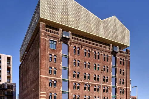 Pakhuis Santos (Nederlands Fotomuseum Rotterdam): historic red-brick warehouse facade crowned by a modern gold-mesh rooftop extension against a clear blue sky.