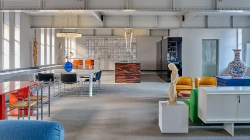 A photo of a room with lots of concrete, colored chairs, tables, a statue and a large vase.
