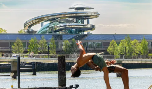 Man in green shorts arches into a backbend on the waterfront dock at Fenix Katendrecht, with the Fenix Food Factory rooftop’s looping spiral slide rising behind him.