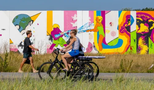Een jonge vrouw op een elektrische fiets met een surfplank achterop en een wandelaar lopen voor een levendige muurschildering met felgekleurde figuren en zomerse watergevechten.