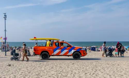 Een opvallende oranje reddingsbrigade-truck rijdt over de strandboulevard, geladen met een surfplank, terwijl gezinnen met kinderwagens en spelende kinderen langs de waterlijn van de Noordzee lopen.