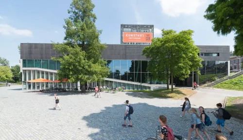Exterior view of Kunsthal Rotterdam, a modern museum building with large windows and an orange sign, surrounded by trees and people walking on the plaza in front.