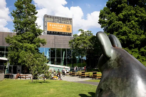 Bronze rabbit sculpture on the lawn before the Kunsthal Rotterdam’s modern glass-and-concrete facade, with visitors near the entrance framed by trees under a partly cloudy sky.