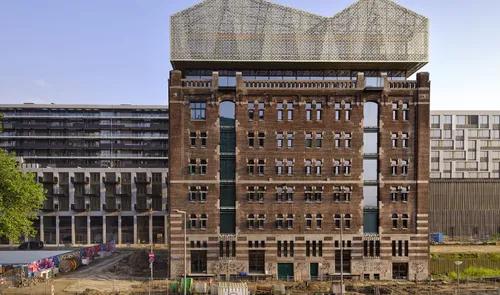 Historic red-brick building facade in Rotterdam with decorative stone window frames, preserved as a standalone front and topped by a modern glass-and-steel rooftop extension.