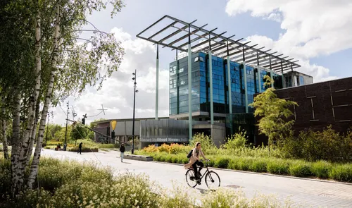 Photo of Nieuwe Instituut Rotterdam exterior: modern glass-and-steel building with an extended canopy, framed by trees and a cyclist riding along the foreground pathway.