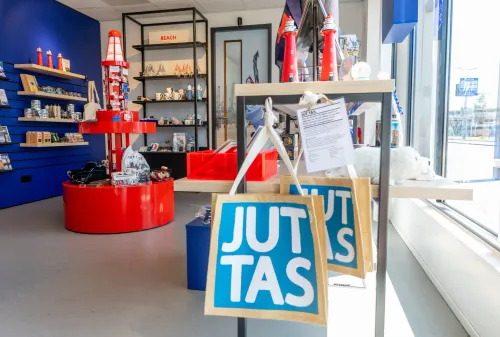 Interieur van de VVV-shop in Hoek van Holland: souvenirrekken met keramiek, ansichtkaarten en een opvallend rood vuurtorendisplay, plus juten tasjes met “JUT TAS”.