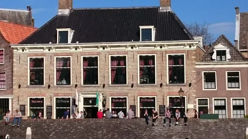 Historic façade of Brouwerij De Pelgrim in Delfshaven, with people sitting along the quay in front of the brick building on a sunny day.