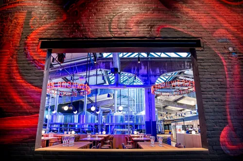 View through a brick opening into Stadshaven Brouwerij, showing brewing tanks, wooden tables, and red neon “Brouwerij” signage.
