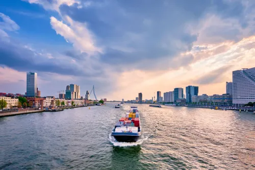 Cargo ship carrying colorful containers sailing on the river Maas with Rotterdam skyline in the background.