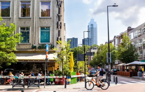Street scene in Rotterdam’s Witte de Withstraat with people dining at outdoor cafés, a cyclist riding by, and modern high-rises visible in the background.