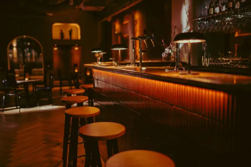 Close-up of a warmly lit bar counter with wooden stools, brass beer taps, and soft amber lighting in De Kunstfabriek.