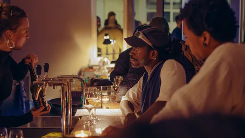 Barkeeper in gesprek met gasten aan een sfeervol verlichte bar, met kaarsen, wijnglazen en warme ambiance op de achtergrond.