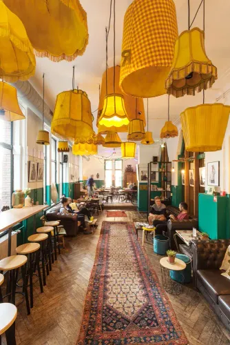 Cozy common area at Hostel ROOM Rotterdam, with vintage decor, patterned rugs, and a ceiling full of mismatched yellow lampshades.