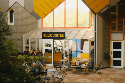 Exterior of KUUB Coffee nestled under the iconic Cube Houses in Rotterdam, with terrace seating and bright window accents.