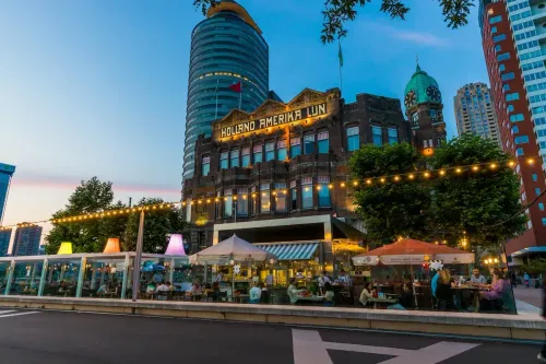 Evening view of Hotel New York with illuminated “Holland Amerika Lijn” signage and a lively outdoor terrace under string lights.