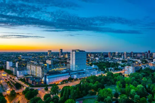 Skyline of Rotterdam at sunset with high-rise buildings, green parks, and city lights starting to glow.