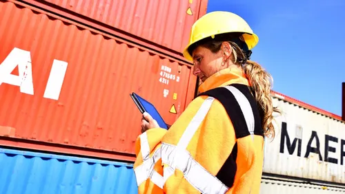 Photo of a woman wearing a safety helmet and checking a tablet computer in the port area.